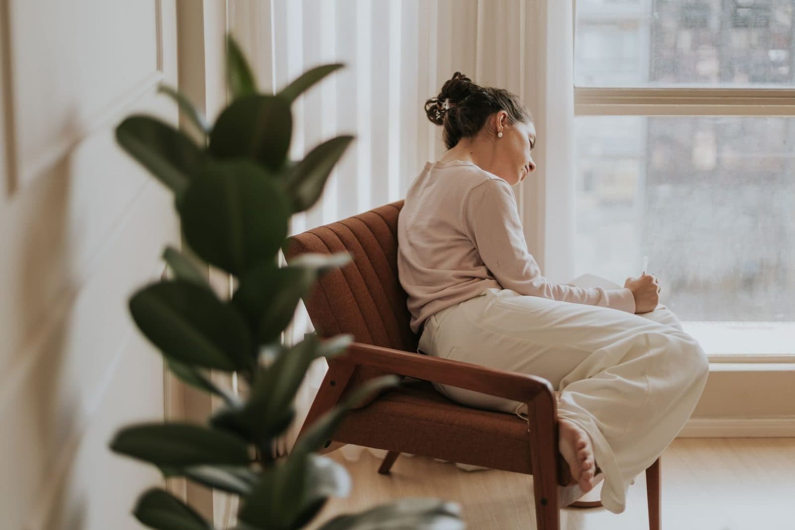 A woman sitting in a chair by a bright window, writing in a notebook, next to a leafy plant
